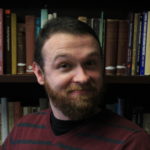 A bearded white man gives the camera a quizzical look. He wears a red sweater and stands in front of a bookcase