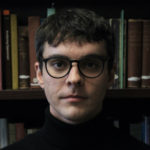 A white man with dark hair stands in front of a bookcase, looking directly into the camera. He wears black glasses and a black turtleneck