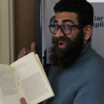 A man is smiling, holding a book open in front of a door. He has dark brown hair and beard, and is wearing glasses and a blue turtleneck.