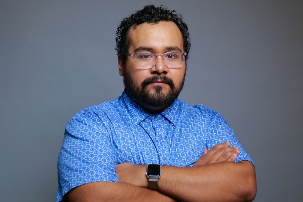 Portrait of a man with curly dark hair and a dark beard wearing clear eyeglasses and a blue button-down shirt.
