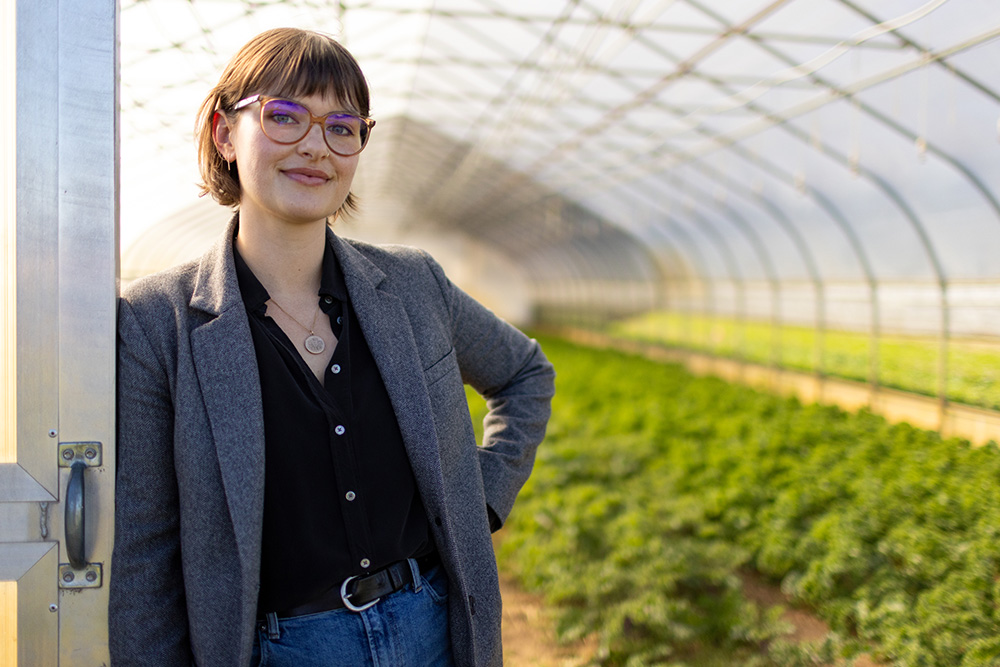 MSU Assistant Professor Megan Dean wearing a blazer and glasses leans against the doorframe of a hoop house. Behind her are leafy green vegetables growing in rows.