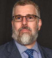 publicity photo of man with grey beard, wearing glasses, blue suit and tie