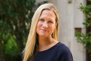 A white woman with straight blonde hair is smiling at the camera and wearing a blue shirt