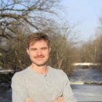 A white man stands in front of a river with arms crossed. He has brown hair and a mustache, and wears a gray shirt