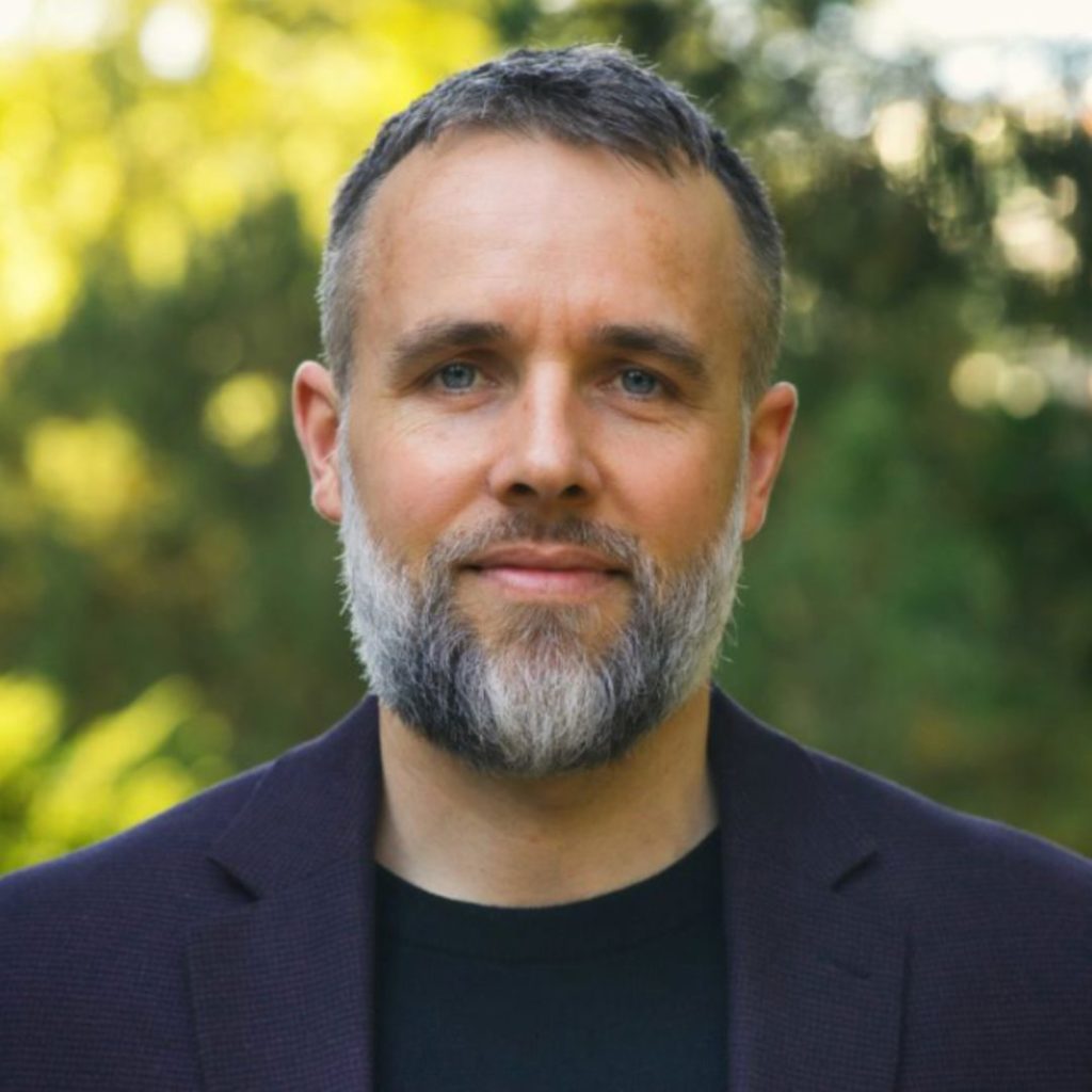 An outdoor portrait of a man with dark, graying hair and beard wearing a navy blazer over a black top.