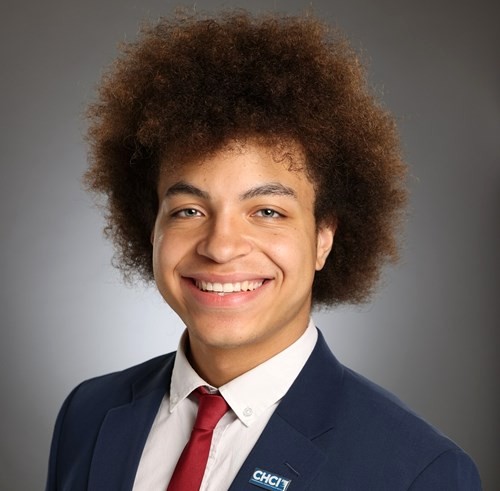 Headshot photo of a man with an afro, smiling, wearing a blue a blue suit, white button-up shirt, and red tie. 