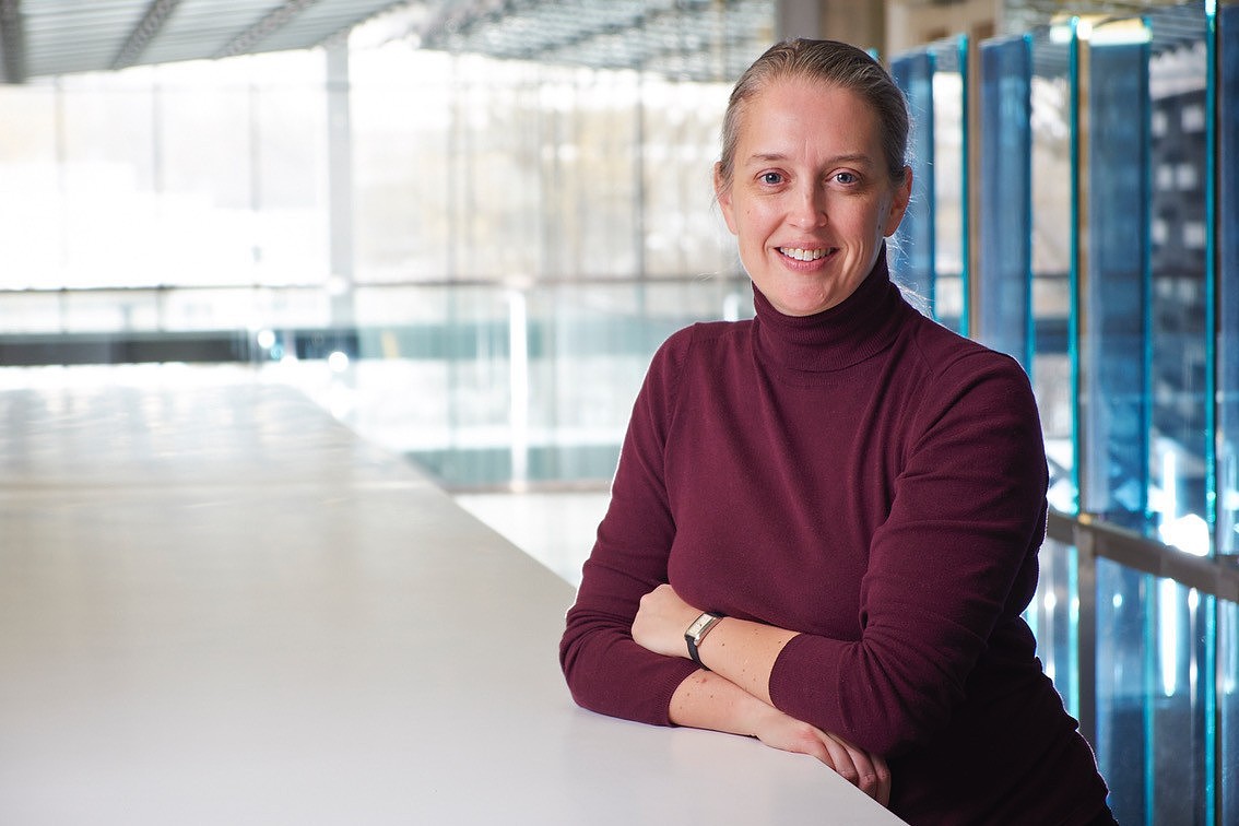 Portrait of a woman wearing a burgundy turtleneck, smiling with her arms folded on a white table in a bright, modern interior with glass walls and blue structural elements in the background.