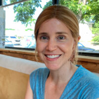 White woman in a blue shirt smiles at the camera. She wears earrings and her brown hair is pulled back.