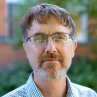A white man with brown hair, a short beard, and glasses looks at the camera.
